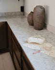 A modern kitchen counter with brown cabinets, stone countertop, two large ceramic vases, a green glass bottle, and a Mushie First Feeding Baby Spoon and small bowl on a pastel polka dot placemat.