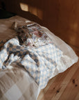 A floral zippered bag and the mushie Organic Cotton Muslin Swaddle Blanket in blue-and-white checks rest on a plaid bedspread, bathed in sunlight on a wooden floor in a cozy, softly lit room.