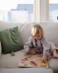 A young child sits on a white couch, assembling the Mushie Wooden Puzzle. Nearby are a green cushion and a small toy, while sunlight streams through large windows—helping develop fine motor skills during play.
