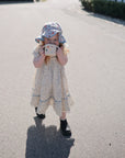 A young girl in a floral dress and sunhat stands on a sunlit pavement, holding the mushie Snack Cup up to her face with both hands. This spill-proof silicone cup is ideal for toddlers. She casts a shadow on the ground while facing the camera.