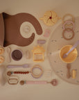 A flat lay of pastel mushie baby feeding and teething items—including bibs, pacifiers, utensils, bowl, cup, teething rings, and a Silicone Placemat—all neatly arranged on a light surface.