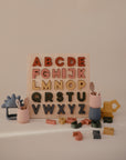 The Mushie Wooden Puzzle, featuring colorful letters, stands upright as an educational toy. Around it, pastel cups, utensils, and vehicles offer engaging ways to develop fine motor skills on a light surface.