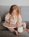 A young red-haired child in a light pink outfit sits barefoot on a gray couch, playing with the mushie Wooden Magnetic Rainbow Maze to build fine motor skills. A white cup sits nearby as she enjoys screen-free play.