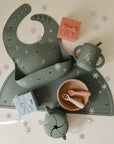 Mushie Silicone Baby Bib in sage green, perfect holiday gift with cups and snowflake confetti.
