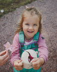 A smiling young girl in turquoise overalls and a pink shirt holds a teapot with flowers as an adult offers her another bloom. Nearby, the mushie Snack Cup—a spill-proof silicone snack cup for toddlers—rests on the gravel path in the grass.