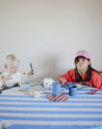A young child with a spoon sits at a table using a mushie Snack Cup, while an older girl in a pink cap enjoys noodles. The blue and white striped tablecloth is covered with various dishes and cups.