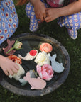 Two kids play with Mushie Whale Bath Stackers in a round water bowl on the grass.
