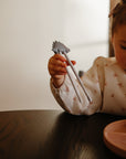 A young child uses mushie Training Chopsticks to pick up noodles from a pink plate.