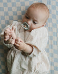 A baby with a pacifier, dressed in a textured cream outfit, lies on a blue and white checkered blanket, holding their feet while reaching for a mushie Unicorn Figurine Teether nearby.