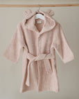 The mushie Bear Robe, a soft, light pink toddler bathrobe made from organic cotton and featuring a hood with bear ears, hangs on a wooden hanger against a neutral background.