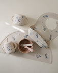 The mushie Snack Cup set for toddlers in a blue floral pattern includes a spill-proof handled cup, lidded silicone snack cup with straw, bib, bowl with two spoons, and placemat, all arranged on a light surface.