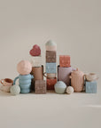 Pastel ceramic cups, teapots, and pitchers are displayed with mushie Mix and Match Present Toys alongside decorative blocks shaped like gift boxes, cupcakes, and spheres, inspiring creative play in a whimsical display.