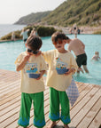 Two young boys in matching yellow shirts and green pants enjoy snacks from mushie Square Dinnerware Bowls by a pool. Both wear sunglasses, one holds a checkered towel, while people swim nearby with hills and the sea in the background.