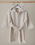 The mushie Bear Robe is a beige organic cotton toddler robe with a hood featuring small bear ears, shown on a white hanger against a neutral background.