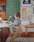 A young child in a mushie Long Sleeve Bib sits in a wooden high chair at a dining table, eating from a bowl with a fork. Green tile and framed art decorate the background, creating a cozy, modern kitchen scene.