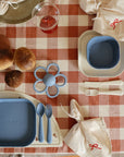 A table for two with mushie Dinnerware Fork and Spoon Set, blue plates and cups on a red and white checkered cloth, round bread rolls, a flower-shaped teether, and ribbon-tied cloth napkins.