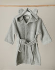 The mushie Bear Robe, a soft, light gray hooded baby robe with cute bear ears, hangs on a wooden hanger against a plain beige background.