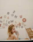 A young girl sits in the tub with her mouth open, while mushie Foam Bath Puzzle pieces stick to the white tiled wall behind her. A wooden bath tray rests across the bathtub.