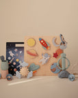 The Mushie Wooden Puzzle features pastel-colored, space-themed pieces like planets, a rocket, astronaut, and UFO to build fine motor skills—shown on a beige surface with a matching background.