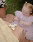 A young girl with red hair in pigtails, wearing a pink dress, sits on a blanket outdoors. She smiles while playing with the mushie Suction Spinner Toy on a cloth, with a pot of pink flowers nearby.