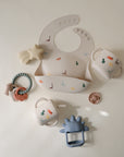 Flat lay of baby items on a light background, featuring a silicone dinosaur-patterned bib, mushie Snack Cup, two handled sippy cups, pale blue teether, beaded animal-shaped teething ring, star-shaped teether, and small cup.