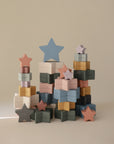 The mushie Nesting Stars Toy features star-shaped blocks in muted blue, pink, green, mustard, and cream for stacking three towers. Inspired by Danish Hygge style, it helps develop fine motor skills.