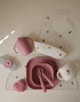 A set of mushie pink and white baby feeding items, including a Snack Cup, bib, plate with fork and spoon, two cups, soft heart accent, and flower-shaped teether—all arranged on a light mat.