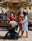 A woman with a mushie Water Resistant Wet Bag and child stand by a stroller near a carousel.