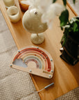 The mushie Wooden Magnetic Rainbow Maze rests on a light wood table by candles, a lamp, and a potted plant atop a soft rug—ideal for screen-free fun and developing fine motor skills.