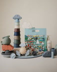 A neatly arranged display features the mushie Reusable Sticker Set, includes stacking cups, cars, a helicopter, sun shapes, puffy stickers, a baby lotion bottle, a feeding spoon, and a play mat with vehicle illustrations.