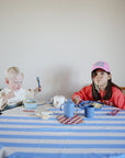 Two kids sit at a table with a blue and white striped tablecloth, eating noodles. They use mushie Dinnerware Cups, while the older child in a pink cap lets noodles dangle from her mouth. A teapot sits nearby.
