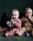Two babies sit on a dark green couch in ribbed pajamas—one pink, one tan—each smiling at the camera while playing with a mushie Robot Rattle Teether.