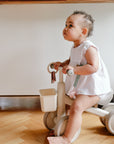 A baby with a mushie Linen Pacifier Clip 2-Pack sits on a tricycle indoors in white clothes.
