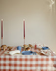 A table with a red and white checkered tablecloth is set with blue dishes, a mushie Dinnerware Fork and Spoon Set, mushrooms, two tall red candles, and other small items against a plain beige wall.