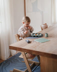 A young child sits at a wooden dining table, tasting food from a muffin tray placed on the Mushie Splat Mat. Sunlight shines through sheer curtains, illuminating a nearby green plant as the focused child explores mealtime.