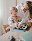 A woman and child sit at a table as the child eats from a food-grade silicone tray. A mushie Silicone Placemat and cup add charm to the wooden table, making this mealtime moment more delightful.