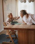 A smiling woman leans over a wooden dining table toward a young child in a high chair offering her food. The child wears the mushie Long Sleeve Bib, which is water resistant. The bright room features modern decor and natural light.