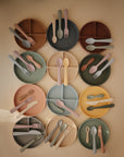 A flat lay of twelve mushie Silicone Suction Plates in soft, muted tones, each with matching forks, spoons, and knives—ideal for stylish baby feeding—arranged neatly on a neutral background.