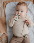 Baby on a mat, wearing beige and chewing on a green mushie Linen Pacifier Clip 2-Pack.