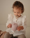A toddler with a FRIGG Daisy Natural Rubber Pacifier holds a mauve sippy cup in white ruffles.
