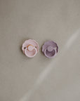 The FRIGG Daisy Silicone Pacifier 2-Pack features one pink and one lavender pacifier with scalloped edges, made from medical-grade silicone for comfort—shown side by side on a neutral, textured background.