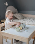 A young child plays at a small table in a cozy bedroom with pastel toy cupcakes and the FRIGG Lucky Symmetrical Silicone Pacifier 2-Pack (0-6 Months), supporting baby oral development. A neatly made bed is visible in the background.