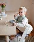 A young child with short blond hair sits in a high chair at a wooden table, eating from a bowl and wearing a light green bib. A mushie Silicone Placemat made from food-grade silicone is on the table amid flowers and toys.