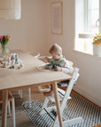 A young child sits in a high chair at a wooden dining table, eating from a bowl with a spoon. The Mushie Splat Mat protects the floor below, while natural light fills the room decorated with flowers and modern minimalist accents.