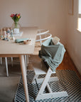 A modern dining room with a light wood table, pastel plates and toys, white chairs, a high chair draped in the mushie Multi-Use Cover (green), and tulips in the background on a checkered mat.