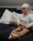 A young child with blond hair sits on a dark couch, wearing a light sweater and brown pants, holding the FRIGG Butterfly Anatomical Silicone Pacifier (6-18 Months); baby cushions and a blanket are visible in the background.