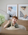 Smiling boy self-feeding with mushie Training Chopsticks at table with art in background.