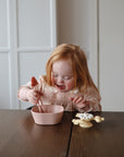 A young girl uses mushie Training Chopsticks to eat from a pink bowl at the table.