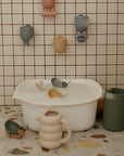 A white tub with Mushie Whale Bath Stackers, toy cups, and a green mug on tiled surfaces.