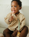 A young child with curly hair smiles on a couch, wearing a beige patterned outfit and holding a pink silicone teether. Attached to their clothes is the mushie Linen Pacifier Clip 2-Pack, featuring a durable silicone clasp.